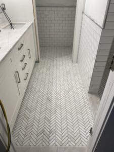 Bathroom with white herringbone floor tiles, complemented by subway tile walls and a sleek white vanity, creating a clean and classic design.