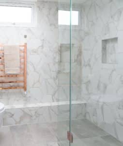 Modern shower with large hexagonal marble tiles, a frameless glass door, and built-in wall niches, complemented by a wooden towel rack.