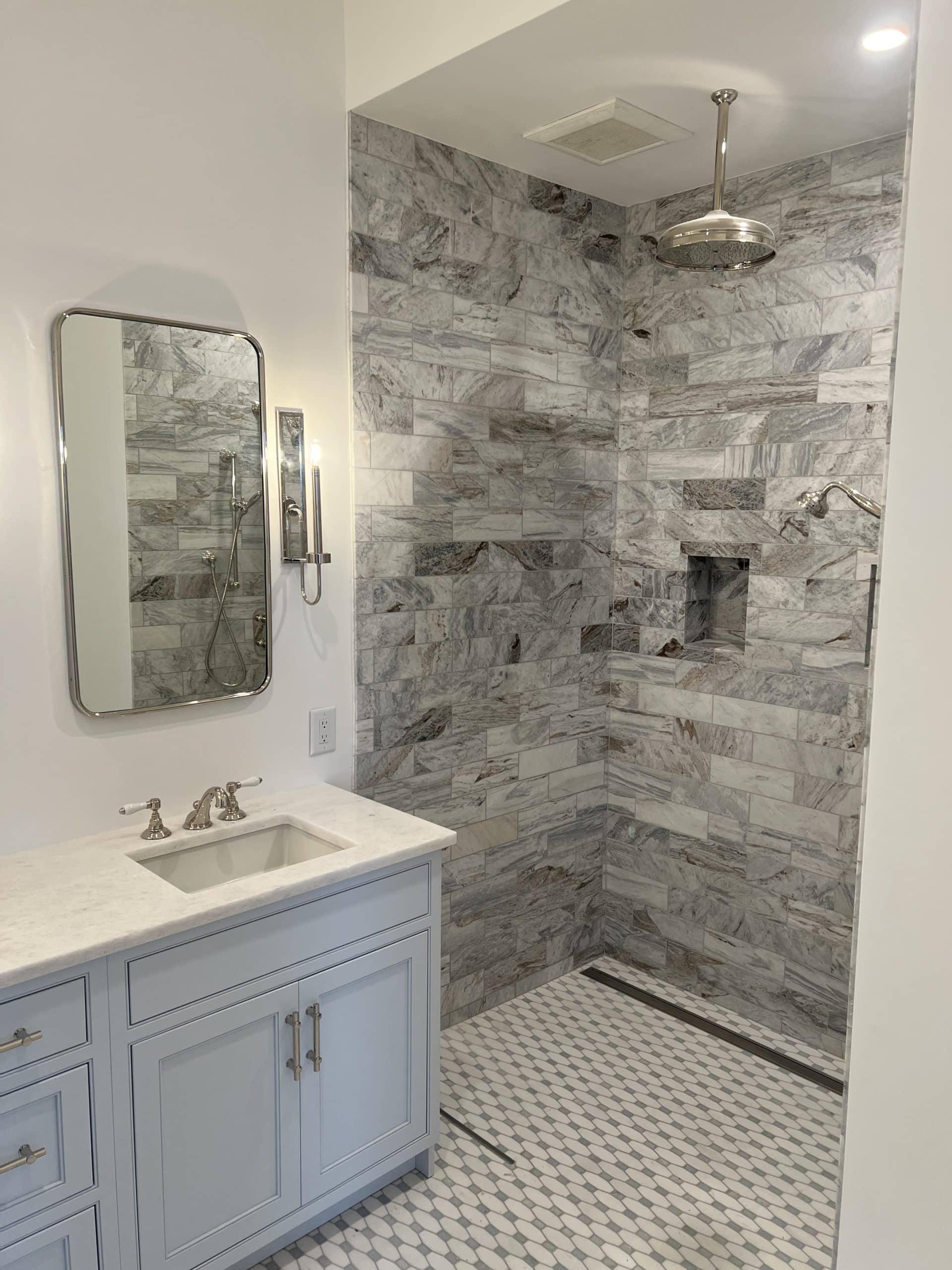 A luxurious bathroom featuring a vanity with a light blue cabinet and marble countertop, an elegant framed mirror, and a spacious walk-in shower with tile walls and floor in various patterns. The shower includes a built-in shelf and a large rain shower head.